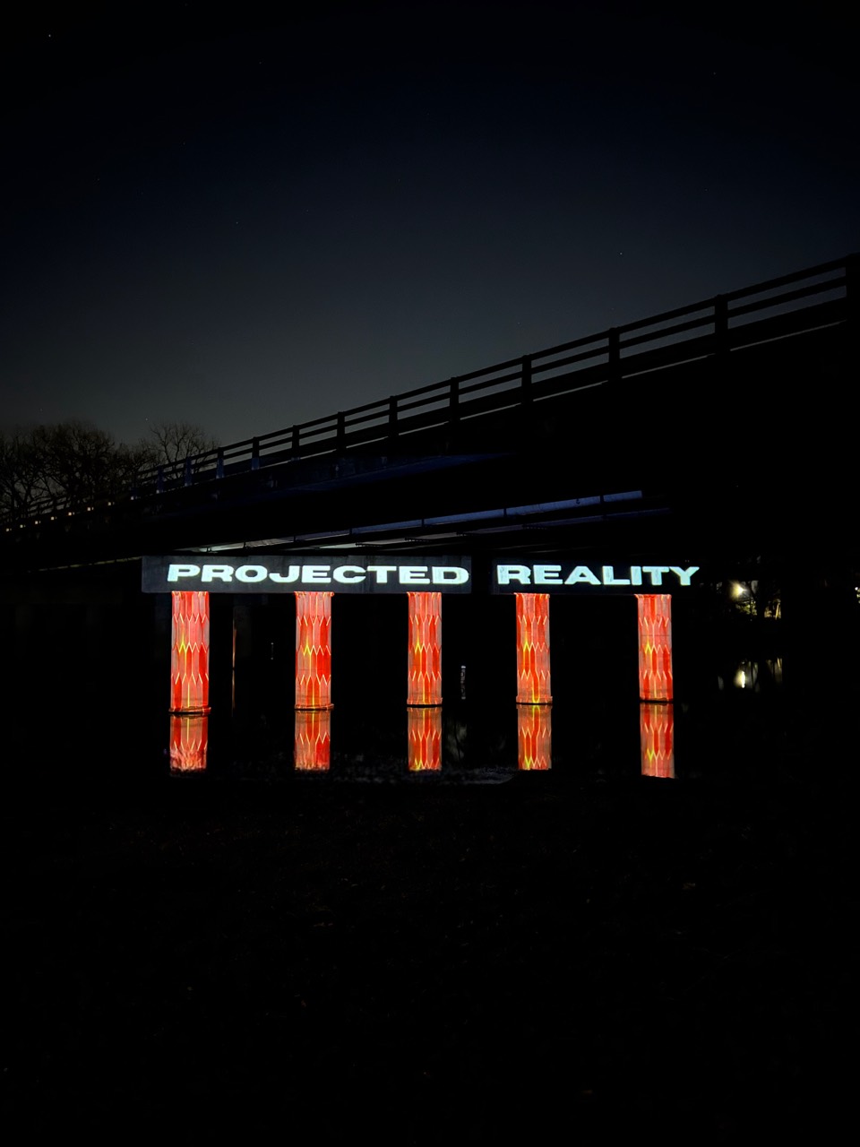 Projected Reality branding and illuminated columns beneath a bridge at night